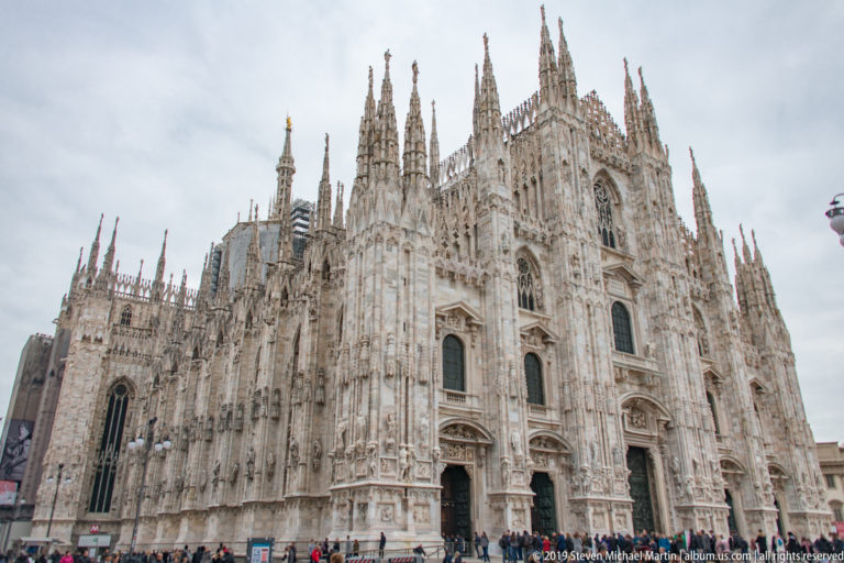 Wide Piazza del Duomo view of Milan Cathedral’s west front and side elevation with visitors gathered at ground level.
