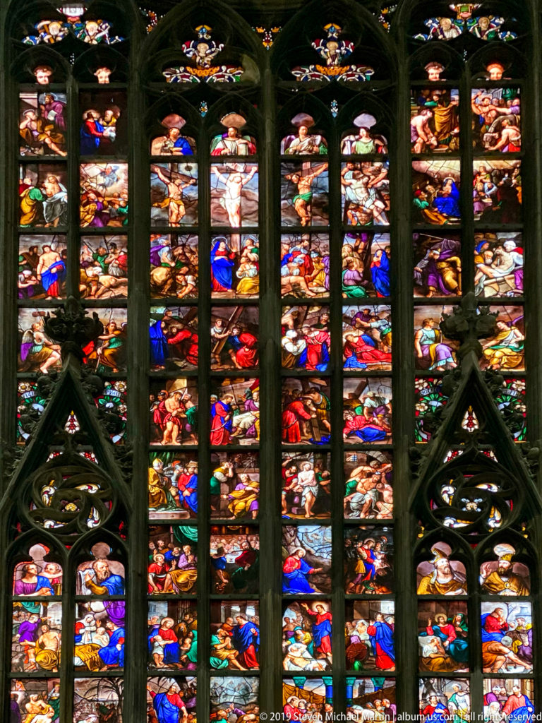 Large Gothic stained-glass window filled with many colorful narrative scenes in tall vertical lights beneath dark tracery.