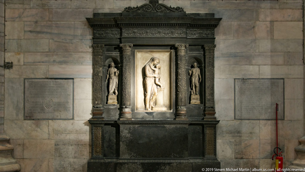 Dark marble wall monument with pale central standing figure, two side statues, columns, and broad rectangular base.