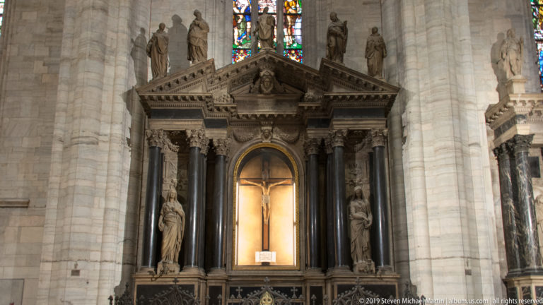 Tall side altar with dark columns, flanking statues, triangular pediment, and brightly illuminated cross in the central niche.