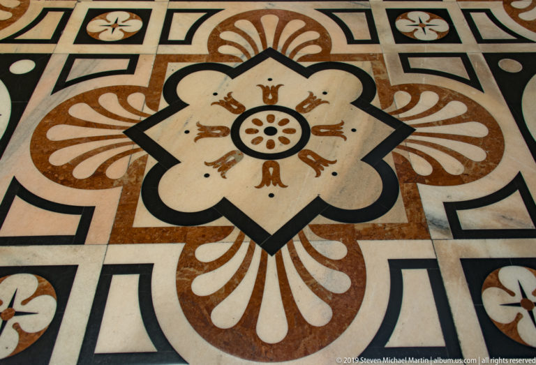 Polychrome marble floor with central rosette, cream petals, rust-brown flourishes, and black geometric borders.