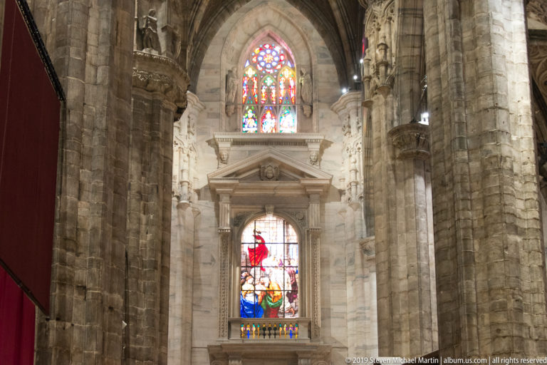 Two stained-glass windows seen between massive pale stone piers and arches inside the soaring cathedral interior.