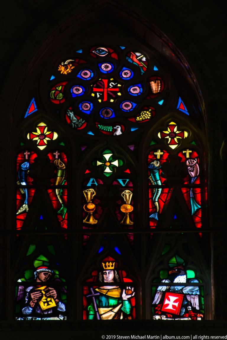 Gothic stained-glass window with standing figures, heraldic emblems, chalice motifs, and colorful circular tracery.
