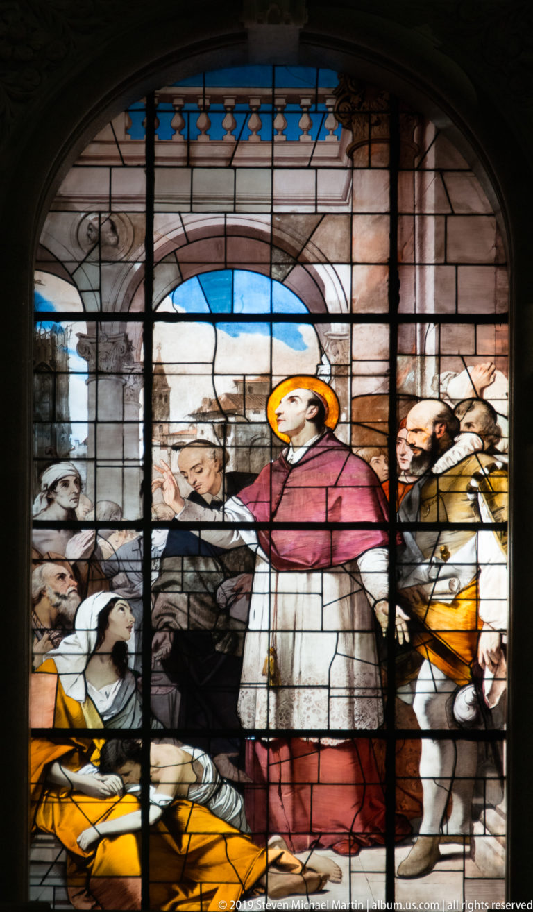Stained-glass scene of a haloed cleric in red and white surrounded by kneeling and standing figures.
