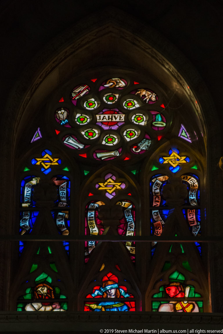 Pointed stained-glass window with circular tracery, heraldic shields, small figures, and geometric colored panels.