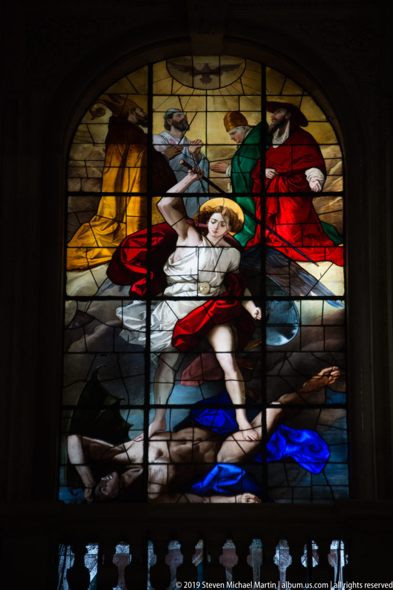 Large stained-glass scene with a central reclining figure surrounded by attendants in red, green, yellow, and blue robes.