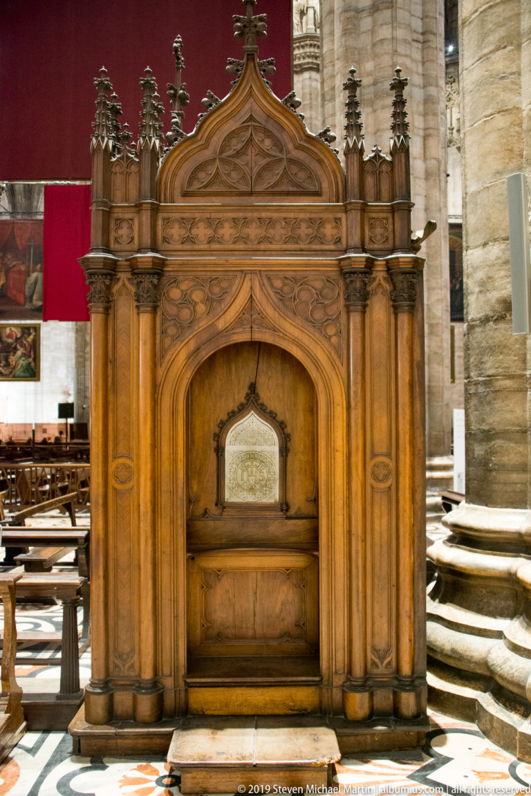 Tall carved wooden ceremonial seat with pointed arch, pinnacles, tracery, and an inset panel on patterned marble flooring.