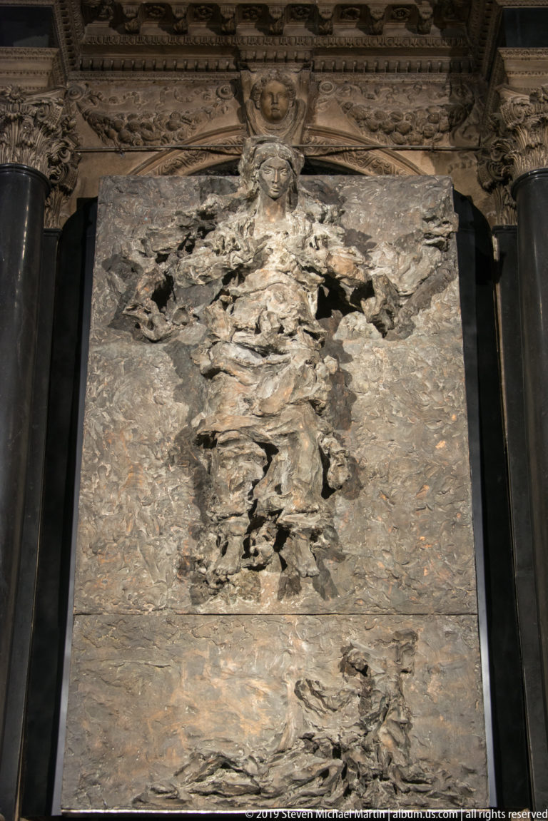 Large weathered stone slab with a damaged central carved design, surface losses, cracks, and mottled gray-brown texture.