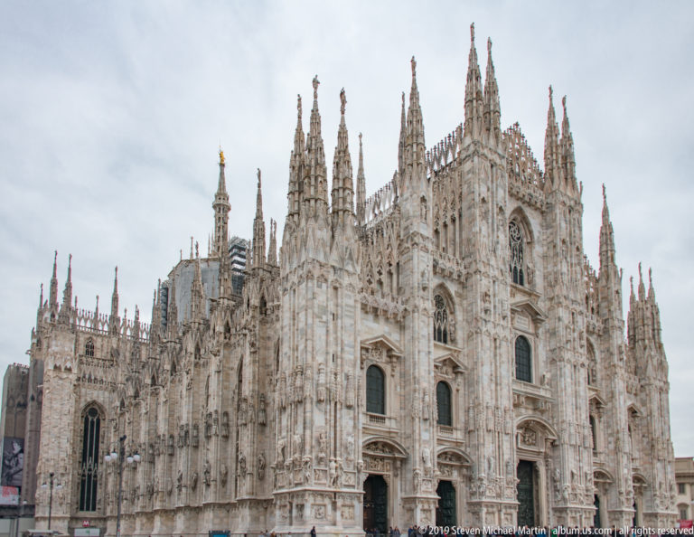 Three-quarter exterior view of Milan Cathedral showing its façade, long side wall, pointed windows, and dense spired roofline.