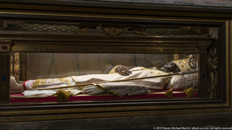 Reclining vested clerical figure displayed behind glass with a pastoral staff, white-and-gold garments, and red lining.