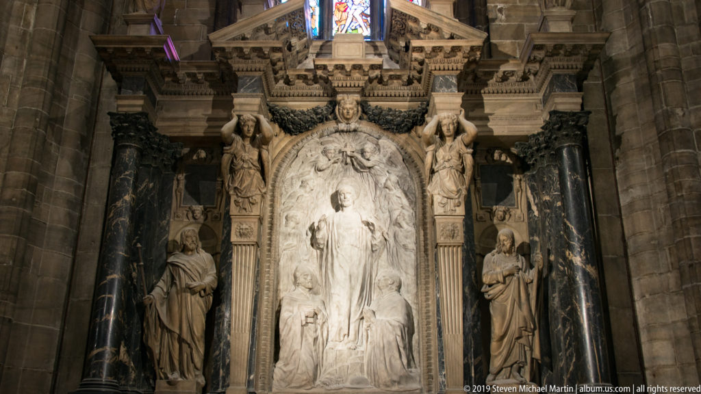 Ornate marble monument with pale central relief, dark columns, standing statues, and carved architectural details.