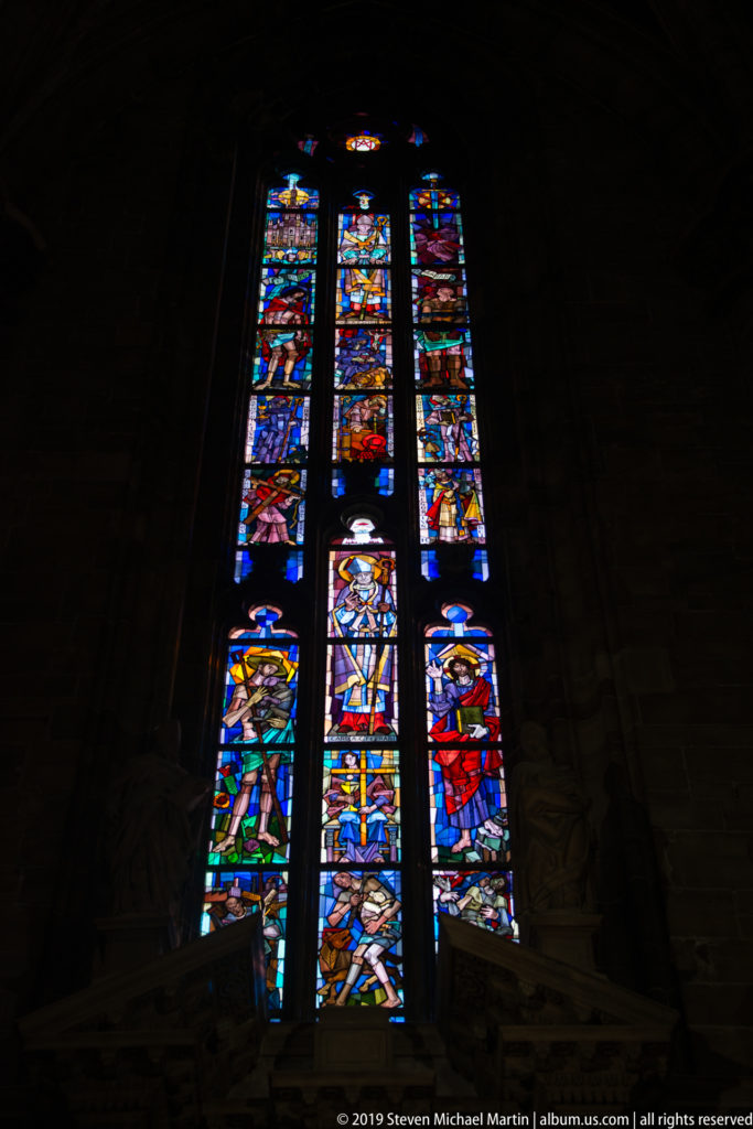 Tall stained-glass window with colorful figures and panels glowing against the dark interior of Milan Cathedral.