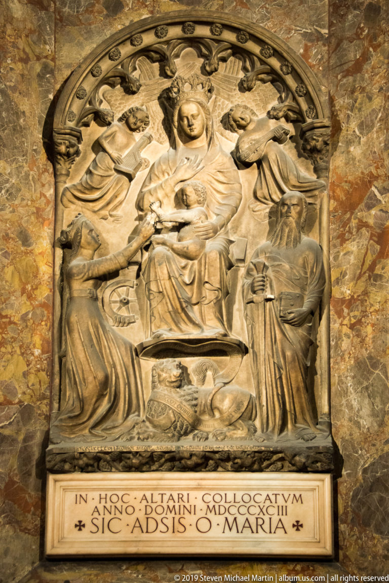 Carved marble memorial relief with a seated clerical figure, attendants, arched ornament, and Latin inscription below.