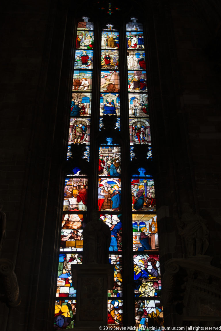 Narrow multi-lancet stained-glass window glowing with many small narrative panels in a dark stone cathedral bay.