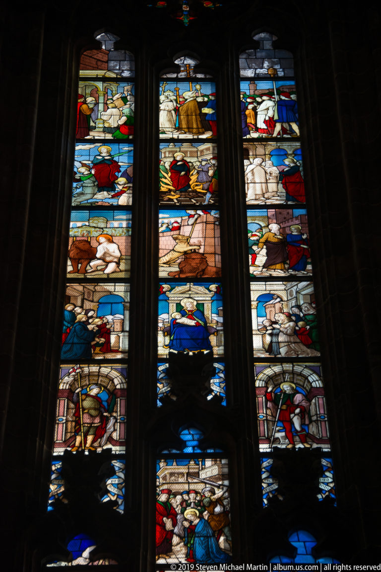 Three tall stained-glass lancets containing stacked biblical or devotional scenes in vivid colors inside the cathedral.