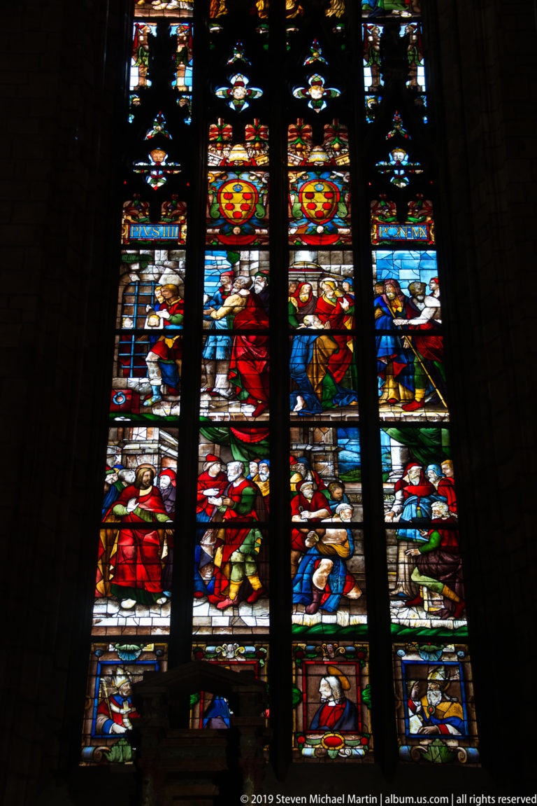 Tall stained-glass window divided by dark mullions into narrative panels of colorful figures and heraldic motifs.