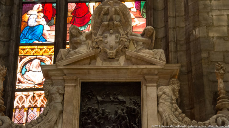 Ornate marble wall monument with a heraldic crest, dark central relief, carved figures, and stained glass glowing behind it.