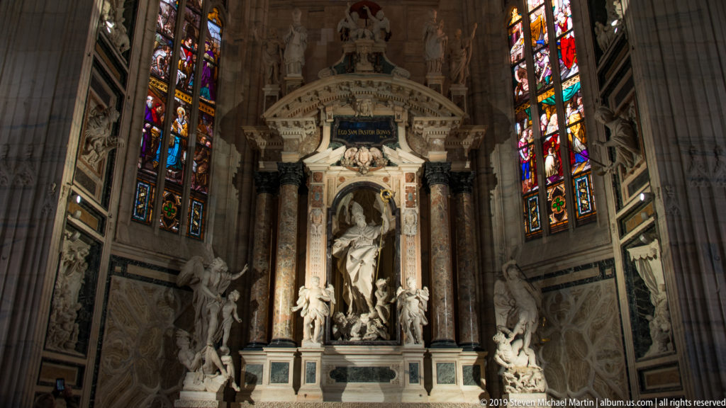 Marble side-chapel altar with central sculpture, dark columns, flanking figures, and stained-glass windows inside Milan Cathedral.