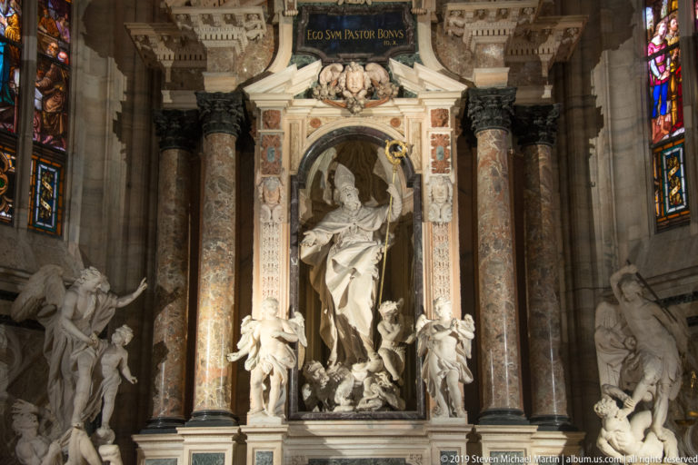 Tall marble memorial with a seated central figure in an arched niche, flanked by angels, colored columns, and sculpture.