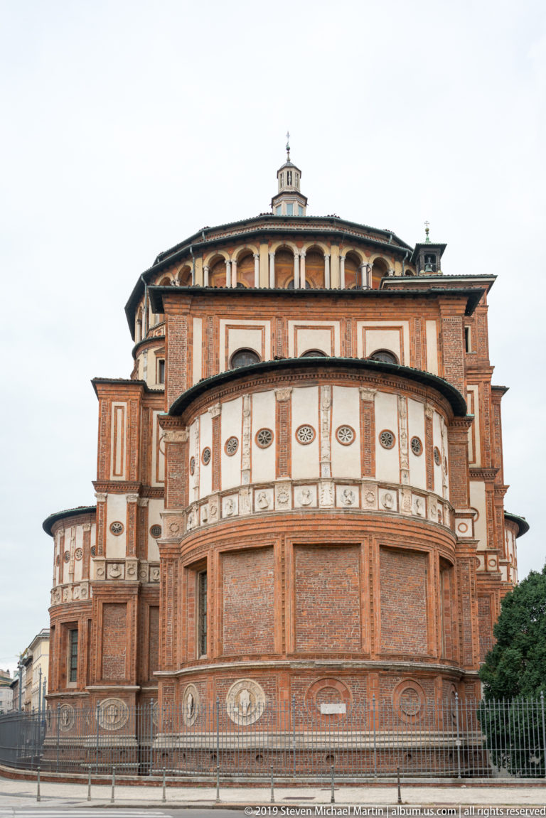 Rear exterior of the Santa Maria delle Grazie tribune with layered brick volumes, pale panels, medallions, and arcaded upper drum.