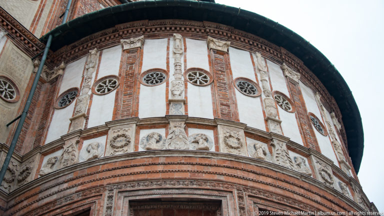 Low-angle view along the rounded tribune, showing circular windows, brick pilasters, pale panels, sculpted frieze, and projecting roof.