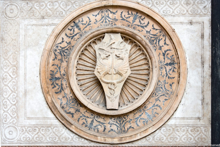 Crowned heraldic shield with an intertwined central device inside a round medallion bordered by blue foliage on a pale wall panel.