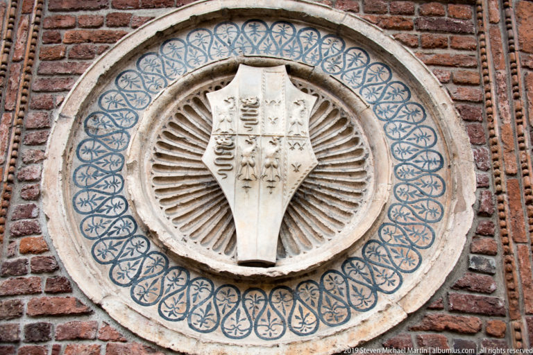 Complex divided shield centered in a circular medallion with radial fluting and blue interlace, set into red brick.