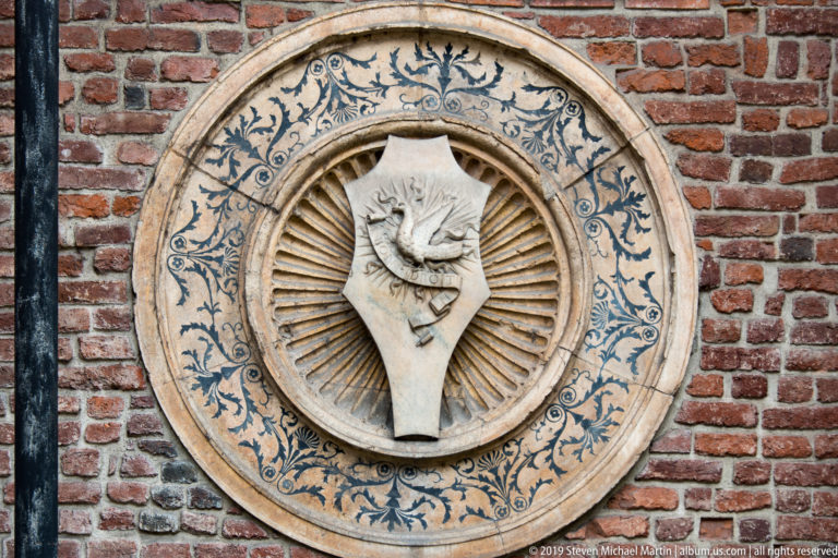 Circular medallion in red brick with a pale shield bearing an animal emblem, surrounded by radial carving and blue foliage.