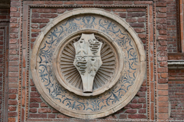 Round heraldic medallion on a brick wall, with a divided shield, radial fluting, and a broad blue ornamental border.