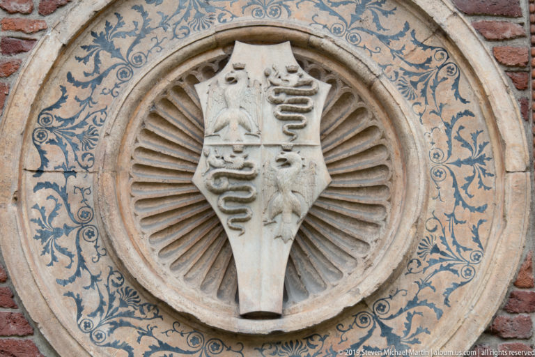 Circular wall medallion with a multi-field heraldic shield, radial carving, and blue scrolling decoration on the Santa Maria delle Grazie tribune.