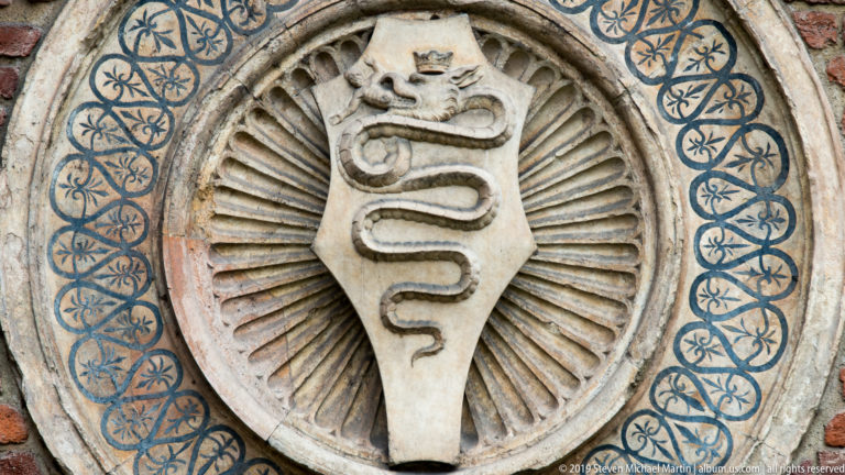 Circular medallion with a shield bearing a coiled serpent, surrounded by radial carving and a blue interlaced border.
