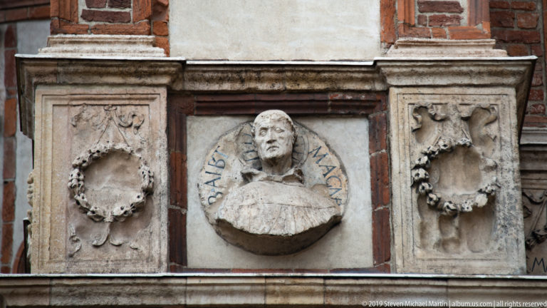 Central sculpted male bust with curved lettering, flanked by two wreath reliefs in the exterior frieze of Santa Maria delle Grazie.