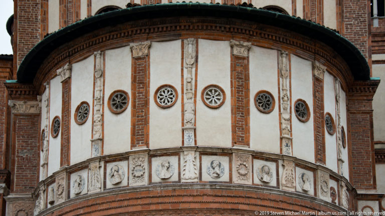 Curved Renaissance tribune exterior with red-brick pilasters, pale panels, circular windows, and a sculpted frieze of roundels and wreaths.