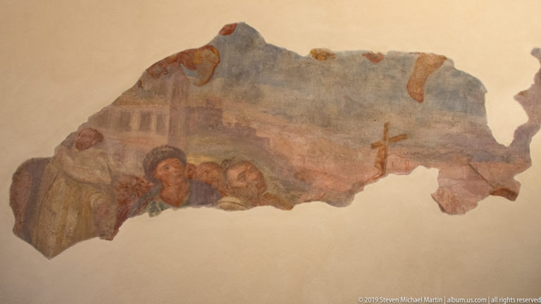 Irregular mural fragment with three figures, faded blue background, reddish architecture, and a small cross at right.