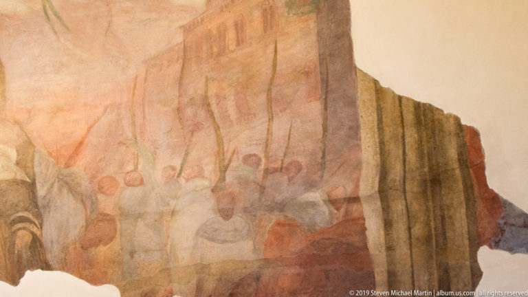 Large fragmentary mural with seated figures, reddish architecture, and tall vertical elements against a pale plaster wall.