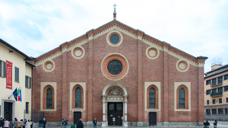 Frontal red-brick façade of Santa Maria delle Grazie with central oculus, smaller round openings, pointed windows, and pale stone portal.