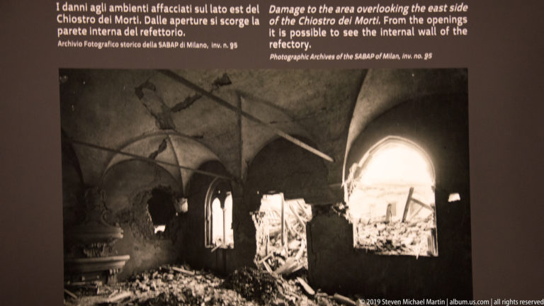 Black-and-white interior view of bomb-damaged cloister vaults with breached walls, rubble, and bright openings to the outside.