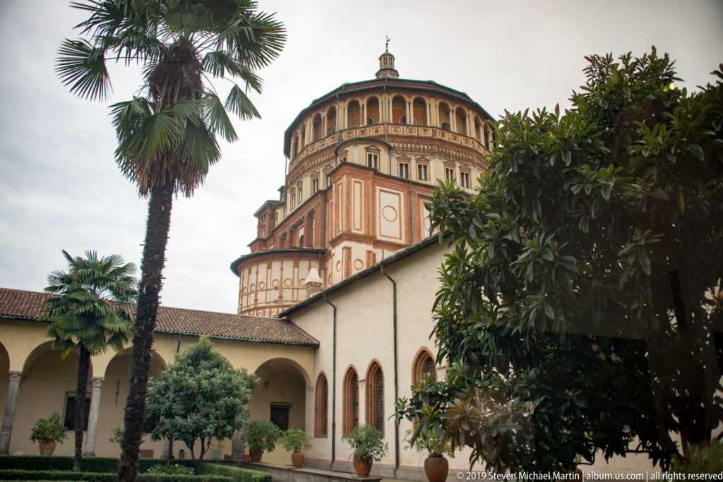 Santa Maria delle Grazie tribune rising above a cloister garden with palms, arcades, red brick, and pale plaster panels.