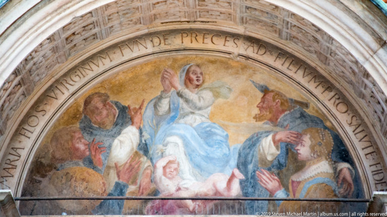 Painted entrance lunette with the Virgin and Child surrounded by kneeling and standing figures, encircled by a Latin inscription.