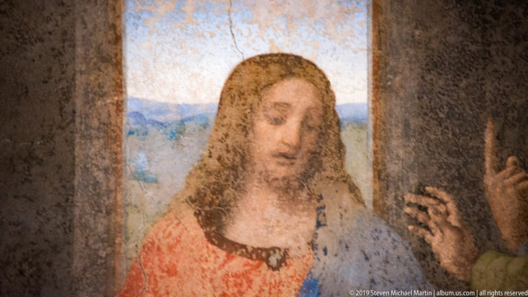 Close portrait of Christ with downcast gaze, red tunic, and blue mantle against the bright central window of The Last Supper.