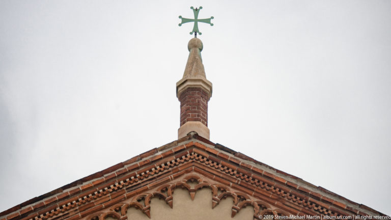 Green metal cross on a stone finial above the red-brick gable and miniature arched cornice of Santa Maria delle Grazie.
