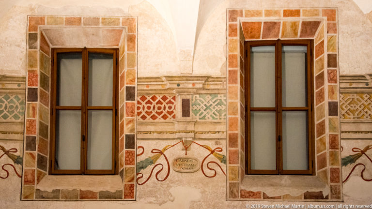Two tall refectory windows framed by faux marble and bands of red, green, and ocher geometric Renaissance decoration.