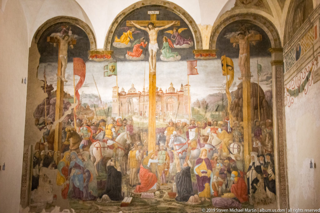 Full 1495 Crucifixion fresco with three crosses above a dense crowd and distant Jerusalem on the refectory wall.