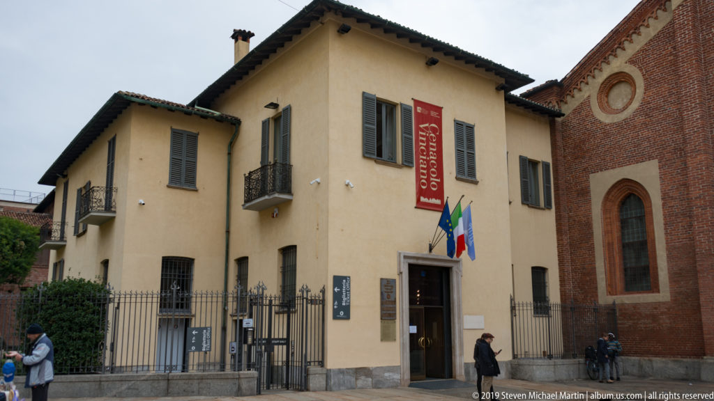 Cenacolo Vinciano entrance in a pale stucco building beside the red-brick wall of Santa Maria delle Grazie, with flags and red banner.