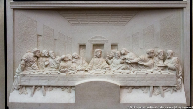 White tactile bas-relief of The Last Supper with raised figures around the table inside a perspectival room.