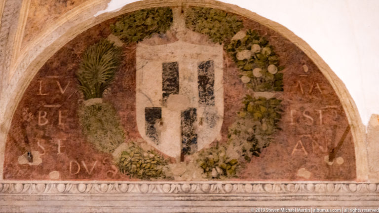 Semicircular painted lunette with a pale heraldic shield, green fruit-and-leaf wreath, faded letters, and classical lower border.