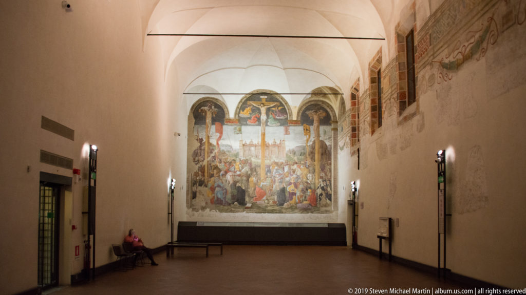 Full view of Montorfano’s Crucifixion at the end of the long vaulted refectory, with a bench and seated visitor in foreground.