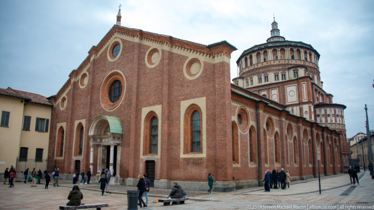 Wide piazza view of Santa Maria delle Grazie showing the red-brick Gothic church and tall Renaissance tribune.