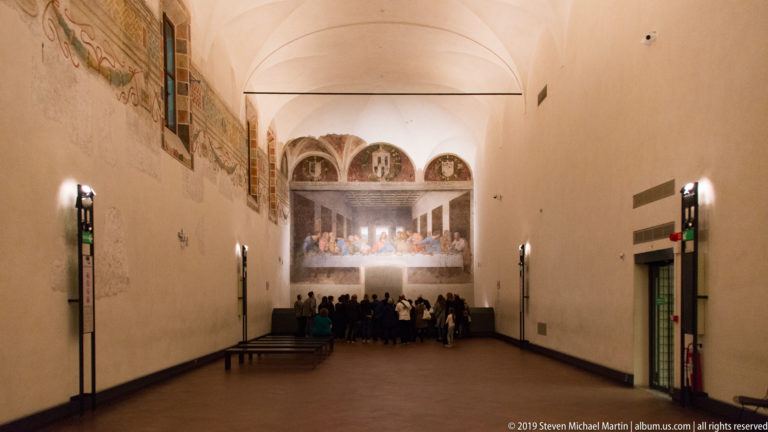 Long vaulted refectory with visitors gathered before The Last Supper on the illuminated far wall.