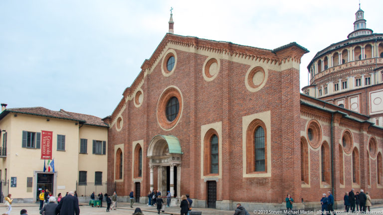 Oblique exterior of Santa Maria delle Grazie with red-brick nave, tall lancet windows, circular oculi, and the Renaissance tribune behind.